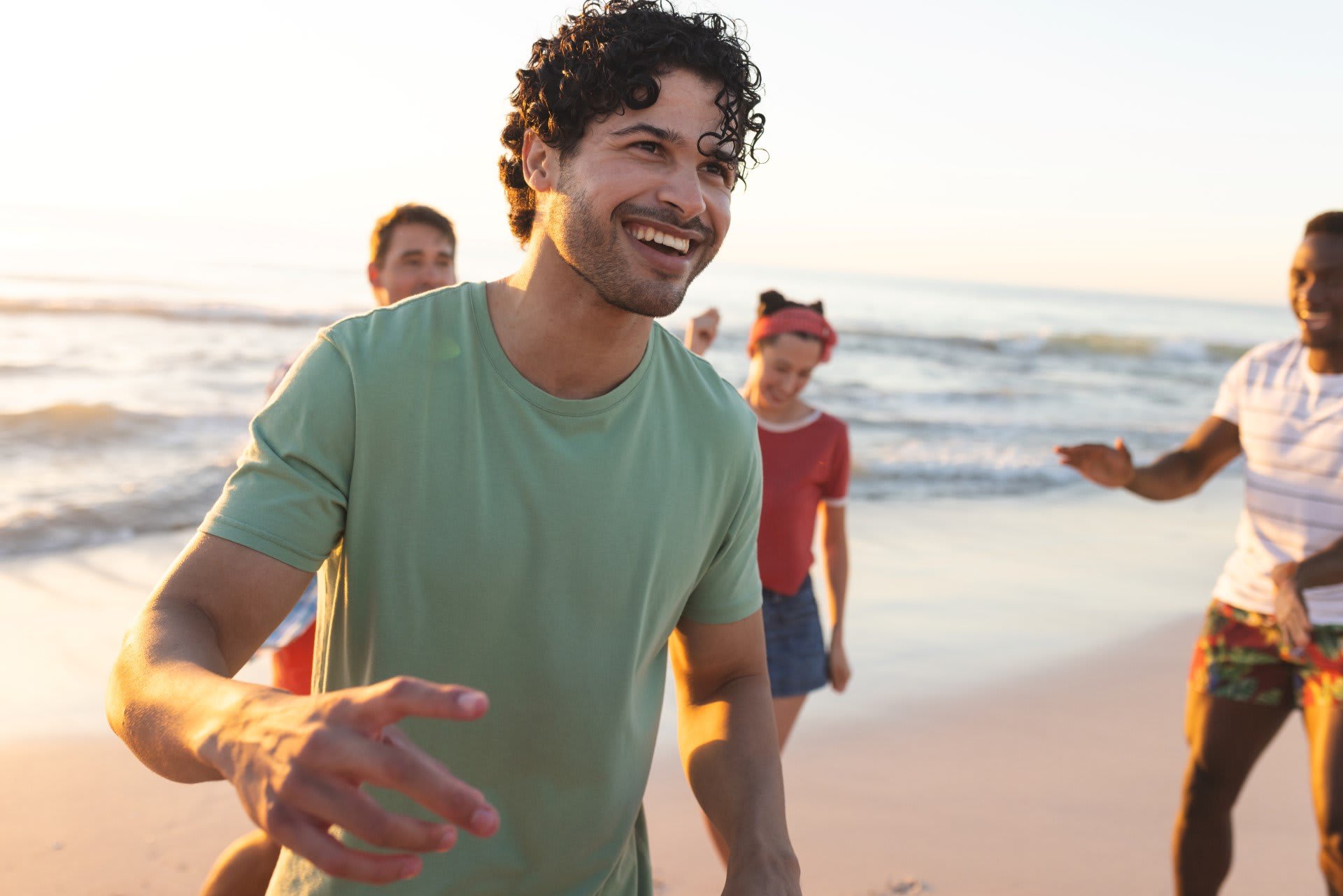 A man in a green shirt smiles and gestures while walking on a beach with three other people in the background. The sun is setting over the ocean.